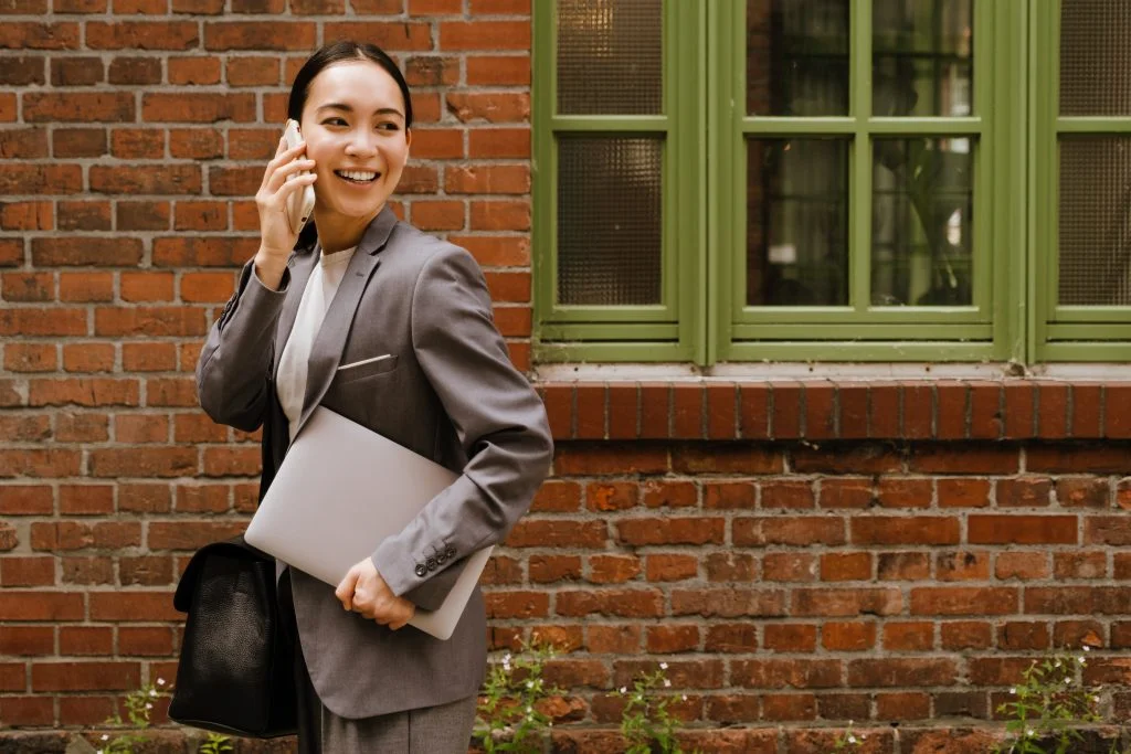 Asian young woman using laptop and talking on cellphone while walking outdoors